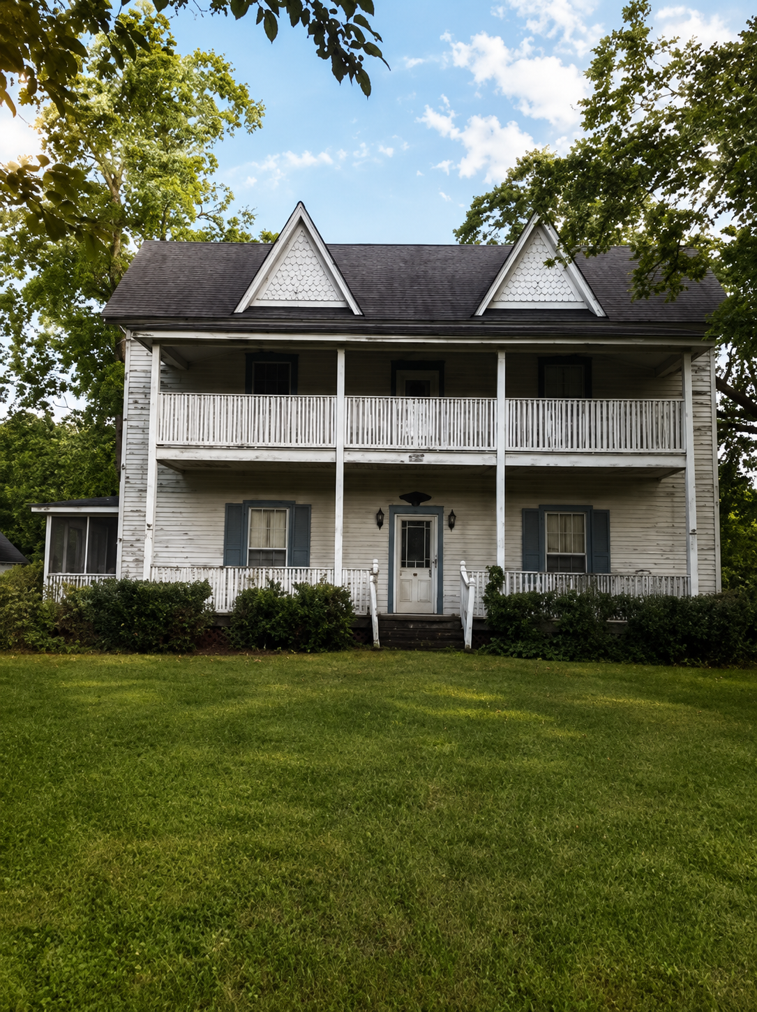 Two-story white farmhouse with black shutters and dual gabled roof surrounded by green lawn and trees